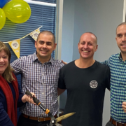 4 people standing together. Person on the left with long hair wearing a black shirt and red scarf holding a pair of giant scissors. Three people standing next to them with heads shaved. Cancer Council decorations in the background
