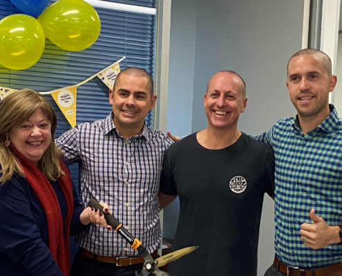 4 people standing together. Person on the left with long hair wearing a black shirt and red scarf holding a pair of giant scissors. Three people standing next to them with heads shaved. Cancer Council decorations in the background