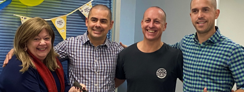 4 people standing together. Person on the left with long hair wearing a black shirt and red scarf holding a pair of giant scissors. Three people standing next to them with heads shaved. Cancer Council decorations in the background