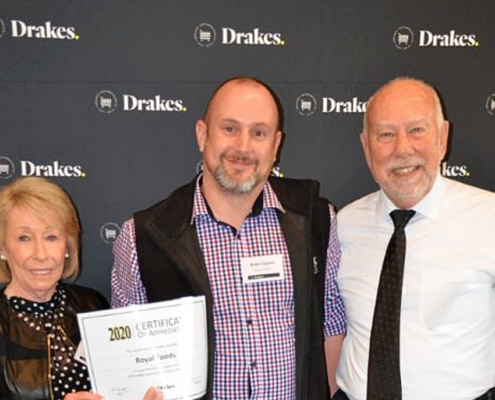 Three people standing together in front of a black "Drakes" backdrop a the Drakes Supermarkets Charity Showbags lunch. Person on the left has a short blonde bob and is wearing a black long sleeve top. Person in the middle is wearing a purple check long sleeve top with a black vest. He is also holding a certificate. Person on the right has a white shirt on and a black tie.
