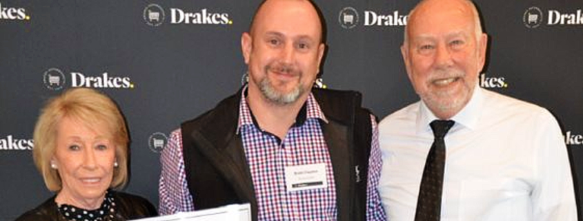 Three people standing together in front of a black "Drakes" backdrop a the Drakes Supermarkets Charity Showbags lunch. Person on the left has a short blonde bob and is wearing a black long sleeve top. Person in the middle is wearing a purple check long sleeve top with a black vest. He is also holding a certificate. Person on the right has a white shirt on and a black tie.