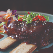 Two Beef Ribs placed in the middle of a blue-grey plate with meet sauce, cut up shallots and red chilli's placed on top. A Mixed cabbage salad can be seen towards the back of the plate.