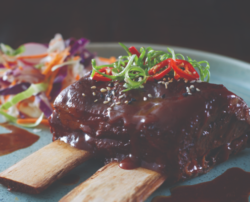 Two Beef Ribs placed in the middle of a blue-grey plate with meet sauce, cut up shallots and red chilli's placed on top. A Mixed cabbage salad can be seen towards the back of the plate.