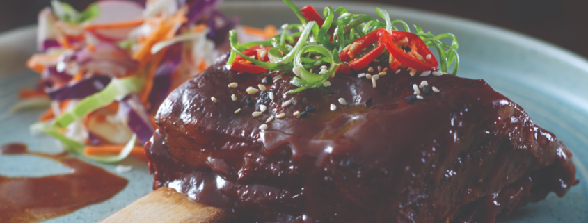 Two Beef Ribs placed in the middle of a blue-grey plate with meet sauce, cut up shallots and red chilli's placed on top. A Mixed cabbage salad can be seen towards the back of the plate.