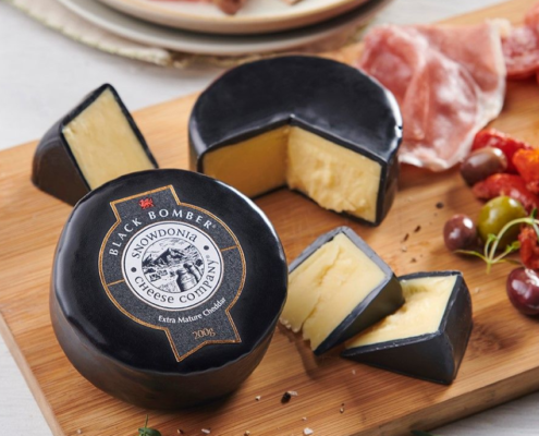 Wooden cheese board with a round of Snowdonia Brie with black wax centered in the picture. Another round of black was brie cut to the right of the board. Cured meats, olives and figs placed artfully around cheese.