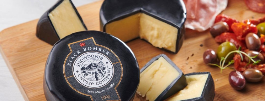 Wooden cheese board with a round of Snowdonia Brie with black wax centered in the picture. Another round of black was brie cut to the right of the board. Cured meats, olives and figs placed artfully around cheese.