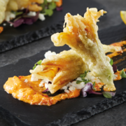 Two fried zucchini flowers in the foreground of the photo, lying on a charcoal serving plate with condiments surrounding. Two more fried zucchini flowers can be seen in the background.