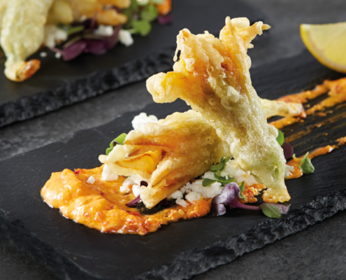 Two fried zucchini flowers in the foreground of the photo, lying on a charcoal serving plate with condiments surrounding. Two more fried zucchini flowers can be seen in the background.