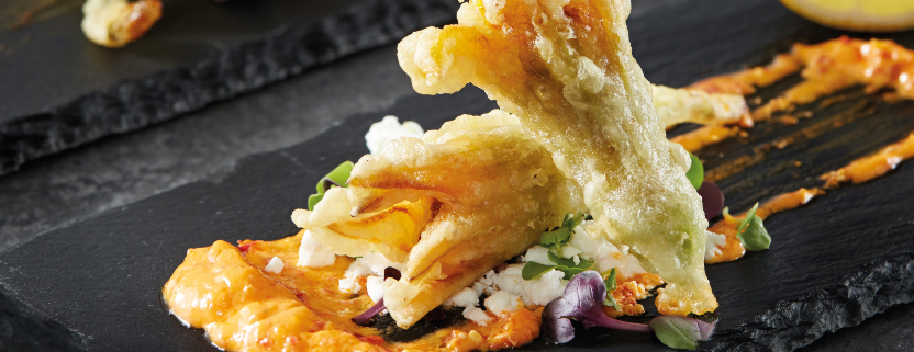 Two fried zucchini flowers in the foreground of the photo, lying on a charcoal serving plate with condiments surrounding. Two more fried zucchini flowers can be seen in the background.