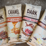 Four packets of Vegan, Plant-based chcolate spread over one another in a small arc. One labelled "Just Creamy", one "Just Caramel", another "Cherry Delight" and the fourth "Just Wh!te"