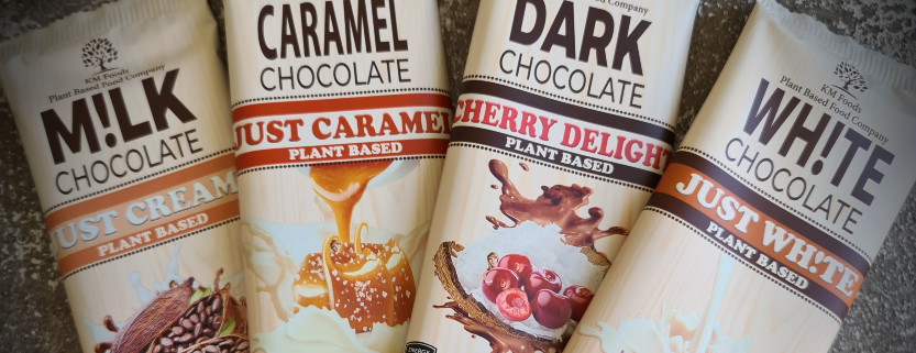 Four packets of Vegan, Plant-based chcolate spread over one another in a small arc. One labelled "Just Creamy", one "Just Caramel", another "Cherry Delight" and the fourth "Just Wh!te"