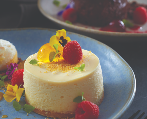Classic Dessert Cheesecake served on a blue plate with a scoop of vanilla ice cream. Flowers and raspberries have dressed the top of the cheesecake