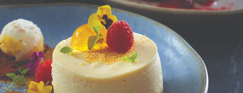 Classic Dessert Cheesecake served on a blue plate with a scoop of vanilla ice cream. Flowers and raspberries have dressed the top of the cheesecake