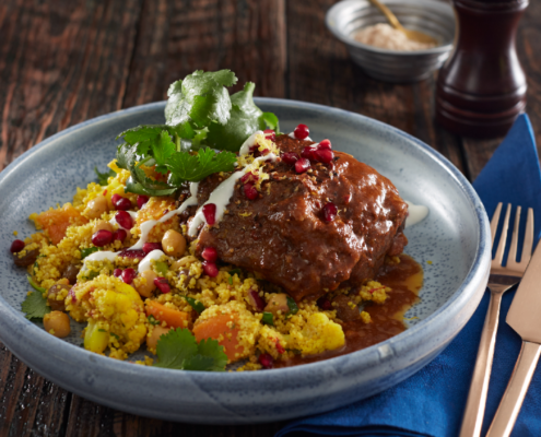 Lamb Shoulder on a light grey plate, sitting on a dark wooden table. Lamb Shoulder is dressed with green leaves & pomengranate seeds, lying on a bed of cous cous